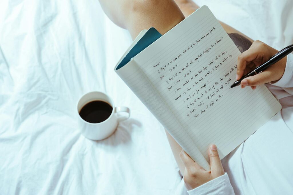 Personne qui écrit dans un cahier, assise sur son lit et une tasse de café à sa gauche.
